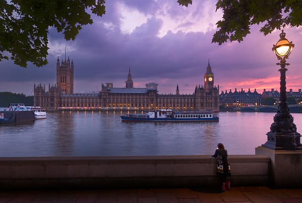 A shot of the Palace of Westminster at sunset, with a boat in the foreground travelling down the River Thames.